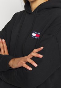 Svart huva gjord av mjukt material, med en framficka och en liten broderad logotyp i blått, vitt och rött på bröstet.