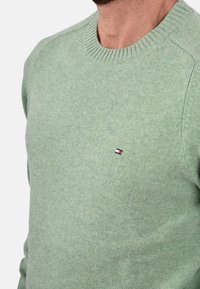 Maglione verde chiaro realizzato in morbido tessuto a maglia, con un colletto a girocollo a coste e un piccolo logo sul petto in rosso, bianco e blu.