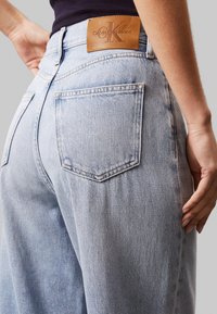 Jeans in denim azzurro chiaro con vestibilità rilassata, dotati di due tasche posteriori e un'etichetta in pelle marrone chiaro con logo impresso sulla vita.
