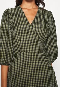 Robe à carreaux verts et noirs avec un design en portefeuille, décolleté en V et manches bouffantes. Le tissu a une apparence texturée.