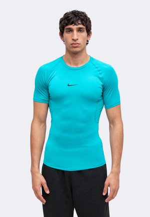 Млад мъж, облечен в прилепнала тюркоазена спортна тениска с къс ръкав на Nike и черни къси панталони, стои срещу обикновен светъл фон.