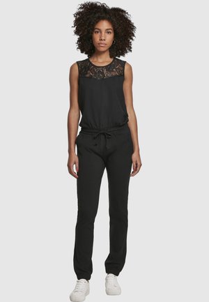 Femme aux cheveux bouclés portant une combinaison noire sans manches avec un détail en dentelle et des baskets blanches, debout devant un fond clair uni.
