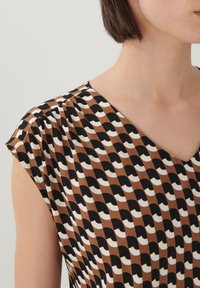 Bruine top met korte mouwen met een golvend zwart-wit patroon. De halslijn heeft een zachte V-vorm, met subtiele plooidetails op de schouders.