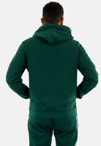 Sudadera de forro polar verde con una gran capucha, puños y dobladillo acanalados, y una textura suave. Mostrada desde atrás, con pantalones a juego.