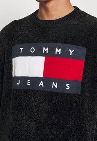 Pull texturé noir avec un grand logo tricoté comprenant un rectangle blanc, des bandes rouges et bleu marine, et le texte "TOMMY JEANS".