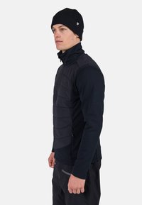 Svart isolerad väst med dragkedja framtill, texturerade paneler och långa ärmar. Bärs över ett svart baslager och kombineras med svarta byxor.
