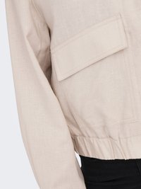 Veste beige à manches longues avec grande poche à rabat rectangulaire et taille élastique portée sur un pantalon noir.