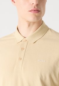 Beige póló, amely színes gallérral, háromgombos záródással és hímzett "HUGO" logóval rendelkezik a bal oldalon. Texturált anyag sima felülettel.
