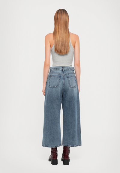 Jeans in denim blu a gamba larga con vita alta, dotati di due tasche posteriori, abbinati a una canotta grigia e stivali borgogna al ginocchio.
