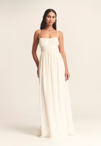 Lange, crème-kleurige jurk met een getailleerde bodice, dunne bandjes en geplooide details bij de halslijn, die overgaat in een losse rok. Zachte textuur.