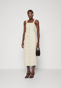 Kreminės spalvos be rankovių midi suknelė su prigludusia liemene, mygtukų uždarymu, šoninėmis kišenėmis ir tiesia sijono dalimi. Derinama su juodais sandalais ir rankine.