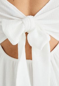 Λευκό ύφασμα με υφή, που διαθέτει λεπτομέρεια με φιόγκο στο πίσω μέρος, αναδεικνύει μια σχέδιο με κοψίματα και ρέουσα σιλουέτα.