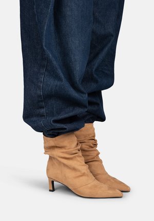 Stivaletti alla caviglia in camoscio marrone chiaro con punte appuntite e tacchi bassi, indossati con jeans blu scuro larghi.