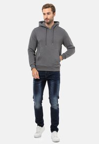 Grauer Baumwoll-Hoodie mit Kängurutasche, verstellbaren Kordeln und gerippten Bündchen, kombiniert mit dunkelblauen Jeans und weißen Sneakern.