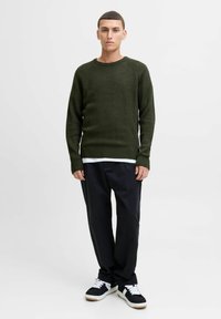 Groene geribbelde gebreide trui met ronde hals, gedragen over een wit shirt, gecombineerd met donkere broeken en zwarte sneakers met witte accenten.