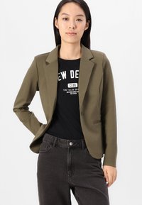 Khaki kavaj med en strukturerad design, enknappstängning och slagkrage, lagd över en svart grafisk t-shirt och mörka jeans.