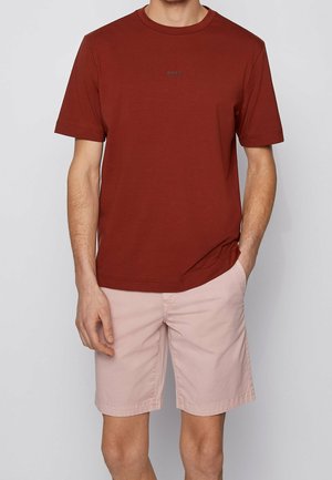 Homme portant un t-shirt à manches courtes couleur rouille et un short rose clair, une main dans la poche, sur un fond gris uni.