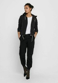 Giacca nera con zip e cappuccio, camicia bianca sotto e pantaloni cargo neri. Stivali trasparenti neri con tacco. Quattro tasche sui pantaloni. Outfit casual.