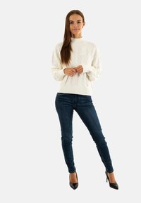 Maglione bianco a collo alto in lavorazione a trecce, abbinato a jeans skinny blu scuro e tacchi alti neri, che mette in risalto i dettagli della maglia lavorata e la silhouette aderente.