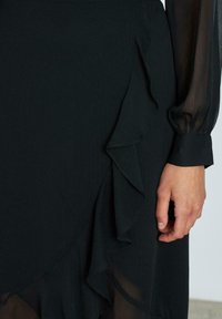 Vestido negro con volantes en capas; hecho de una tela ligera y texturizada. Presenta mangas transparentes y un puño ajustado en la muñeca.