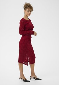 Abito rosso in pizzo a maniche lunghe con una silhoutte aderente e uno spacco laterale. Abbinato a scarpe nere a punta con un tessuto liscio.