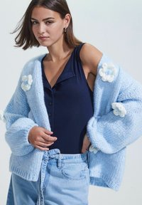 Modèle portant un cardigan en tricot bleu clair avec des accents floraux, un top marine et un jeans en denim, posant avec confiance les mains sur les hanches.