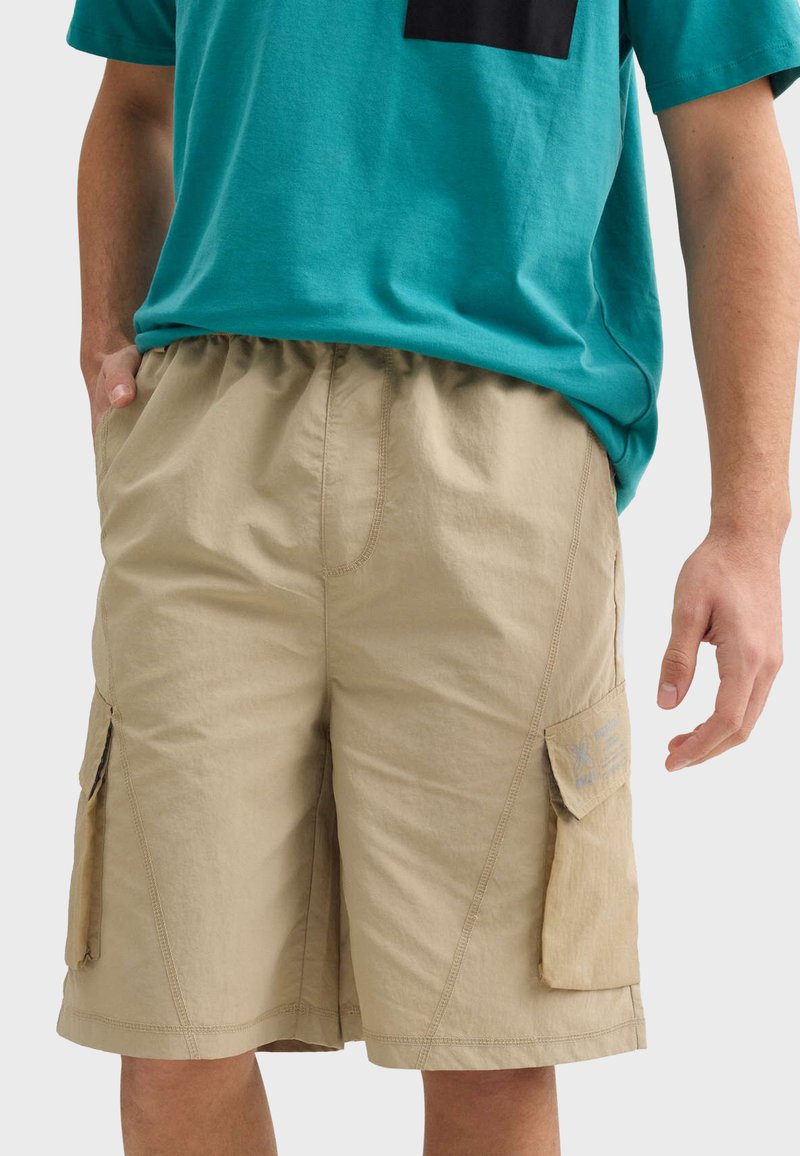 Khaki cargo šortky s elastickým pásom, bočnými vreckami a jemnou textúrou. Obsahuje štepovacie detaily a minimalistický dizajn.