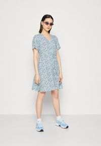 Robe florale bleu clair avec des manches courtes et un décolleté en V, taille cintrée, jupe évasée. Associée à des baskets bleu clair et des lunettes de soleil.