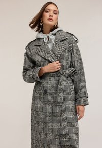 Soprabito a righe bianche e nere con bottoni doppi, vita cintata e ampi rever, indossato sopra un felpa grigia con cappuccio.