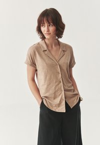 Beige, galléros, rövid ujjú ing, gombos záród�ással és feltekert ujjakkal. Fekete, laza szabású nadrággal párosítva. Simára szőtt anyagtextúra.