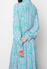 Robe à manches longues avec un motif ondulé turquoise et rose, fabriquée en tissu doux. Les poignets sont ornés de boutons et le design descend jusqu'à l'ourlet.