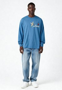 T-shirt bleu à manches longues avec le texte "je t'aime", orné d'un motif de chérubin et de coeur, assorti à un jean bleu clair et des baskets blanches.