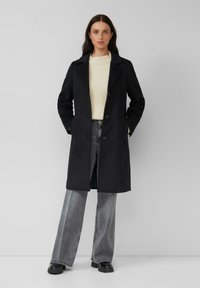 Schwarzer Woll-Mix langer Mantel mit Reverskragen, Knöpfen vorne und Seitentaschen, kombiniert mit einem cremefarbenen Pullover und grauen weit geschnittenen Jeans.