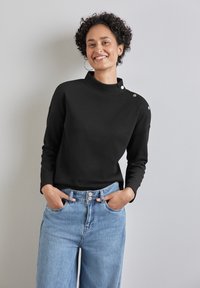 Top nero a costine con scollatura alta e dettagli con bottoni sulla spalla, abbinato a jeans chiari a vita alta. Sfondo luminoso.