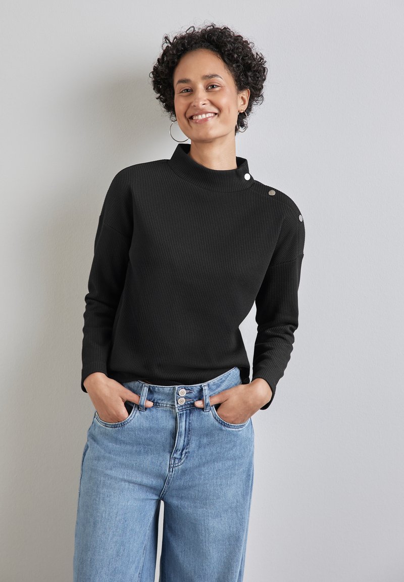 Top nero a costine con scollatura alta e dettagli con bottoni sulla spalla, abbinato a jeans chiari a vita alta. Sfondo luminoso.