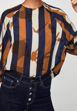 Blouse ample avec des rayures abstraites verticales bleu marine, marron, crème et noir, rentrée dans un jean bleu foncé taille haute avec boutons apparents.