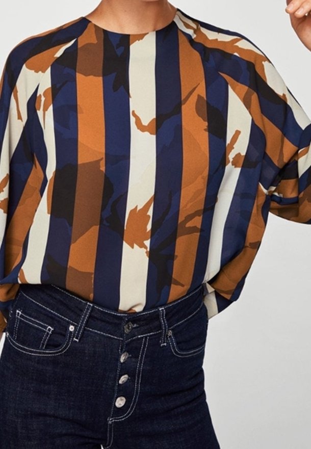 Blouse ample avec des rayures abstraites verticales bleu marine, marron, crème et noir, rentrée dans un jean bleu foncé taille haute avec boutons apparents.