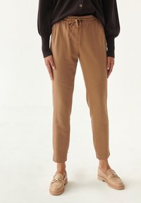 Lysbrune joggerbukser med snøre i taljen, tætsiddende ben og tekstureret stof. Kombineret med beige loafers med guldfarvede hardware-detaljer.