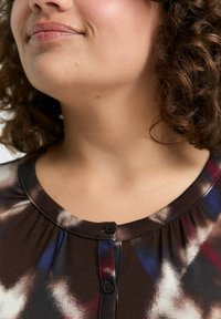 Vrouw met krullend haar, gekleed in een donkere blouse met een patroon en knopen langs de halslijn, zichtbaar van kin tot borst.