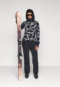 Skiausrüstung mit einer schwarz-weiß gemusterten Jacke mit Kapuze, schwarzen Hosen und schwarzen Skischuhen. Hält einen Ski mit Bindungen.
