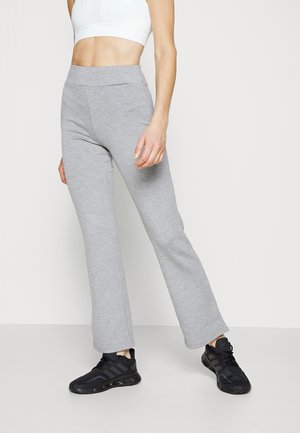 Personne portant un pantalon de survêtement évasé gris clair, un crop top blanc et des chaussures de sport noires, debout sur un fond blanc.