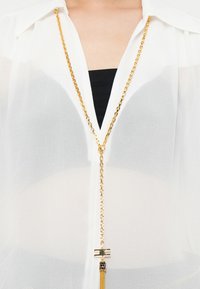 Gold chain necklace with a geometric pendant and tassels worn over a sheer white blouse and black top underneath.