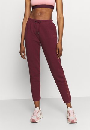 Vrouw draagt bordeauxrode joggingbroek met trekkoord in de taille en roze-witte sneakers, staand tegen een effen witte achtergrond.