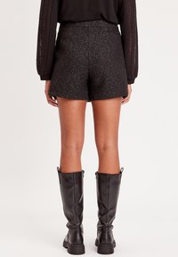 Schwarze strukturierte Shorts mit hoher Taille und dezentem metallischem Faden, kombiniert mit kniehohen schwarzen Lederstiefeln mit seitlichen Reißverschlüssen.