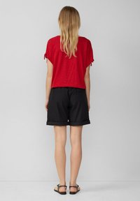 Rode top met korte mouwen en strikaccenten, gecombineerd met opgerolde zwarte shorts. De outfit wordt aangevuld met platte sandalen. Eenvoudig, casual ontwerp.