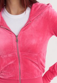 Roza zip-up hoodie izrađena od mekanog, teksturiranog materijala, s prednjim džepom i srebrnim patentnim zatvaračem. Nosila se preko bijelog rebrastog topa.
