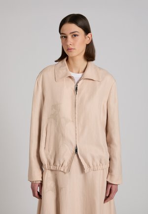 Giovane donna con capelli castano chiaro alla lunghezza delle spalle, indossa una giacca beige con cerniera e una gonna abbinata con un sottile ricamo floreale.