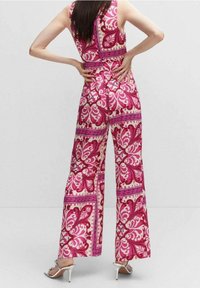 Combinaison colorée avec un motif floral en rose et rouge sur un fond crème, jambes larges et corsage ajusté avec un col montant.