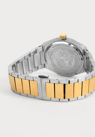 Montre-bracelet en métal argenté et doré avec tête de Méduse gravée au dos et couronne dorée sur fond blanc.