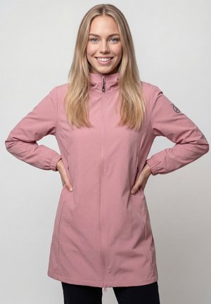 Lächelnde Frau mit langen blonden Haaren, die eine pinke Jacke mit Reißverschluss trägt, Hände in der Hüfte, stehend vor einem einfarbigen hellgrauen Hintergrund.